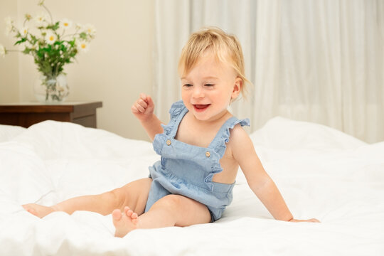 Pretty smiling caucasian blonde baby girl of 1-2 years old sit on white blanket,laugh in bedroom.Cute kid having fun,emotional child,Concept of good sheets or infant before going to bed or wake-up