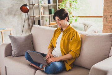 Photo of positive freelancer programmer guy hold laptop hands typing sit sofa wear yellow shirt home indoors