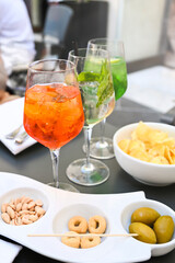 There are three glasses on the table with different colorful cocktails, surrounded by a variety of snacks