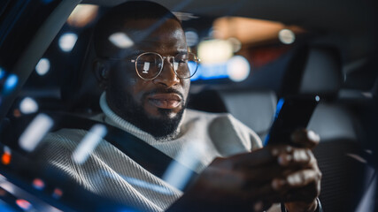 Stylish Black Man in Glasses is Commuting Home in a Backseat of a Taxi at Night. Handsome Male Using Smartphone and Looking Out of Window while in a Car in Urban City Street with Working Neon Signs.