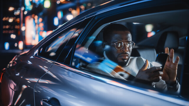 Stylish Black Man In Glasses Is Commuting Home In A Backseat Of A Taxi At Night. Handsome Male Using Smartphone And Looking Out Of Window While In A Car In Urban City Street With Working Neon Signs.