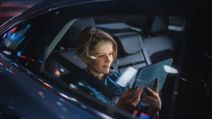 Young Boy is Sitting on Backseat of a Car, Commuting Home at Night. Passenger Watching Funny Entertainment on Tablet Computer while in Taxi in City Street with Working Neon Signs.