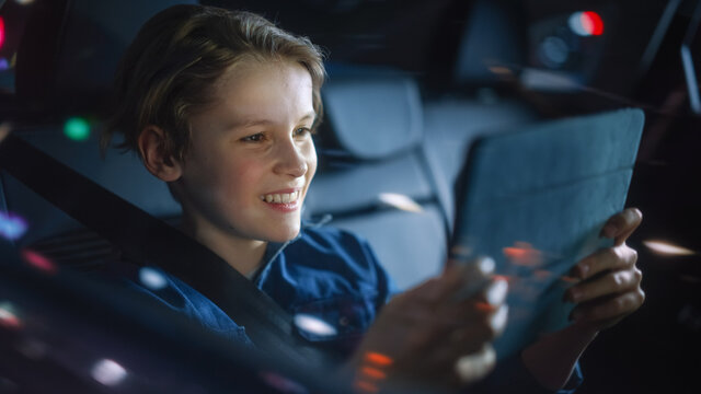 Handsome Young Boy is Sitting on Backseat of a Car, Commuting Home at Night. Passenger Playing Video Game on Tablet Computer while in Taxi in City Street with Working Neon Signs. - Powered by Adobe
