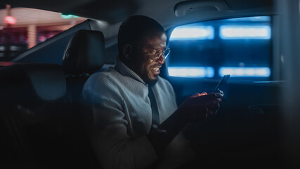 Happy Black Man in Glasses is Commuting Home in a Backseat of Taxi at Night. Handsome Male Using Smartphone and Smiling while in a Car in Urban City Street with Working Neon Signs.