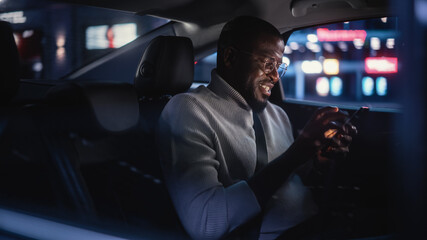 Happy Black Man in Glasses is Commuting Home in a Backseat of Taxi at Night. Handsome Male Using Smartphone and Smiling while in a Car in Urban City Street with Working Neon Signs.