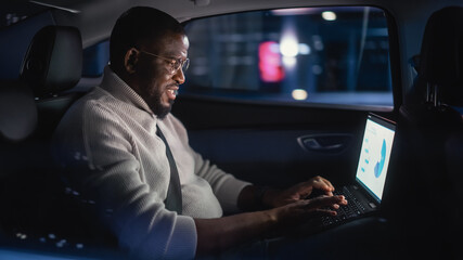 Stylish Black Man in Glasses is Commuting Home in a Backseat of a Taxi at Night. Male Using Laptop...