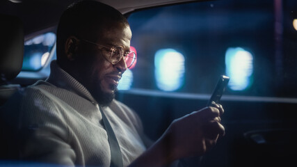 Happy Black Man in Glasses is Commuting Home in a Backseat of Taxi at Night. Handsome Male Using Smartphone and Smiling while in a Car in Urban City Street with Working Neon Signs.