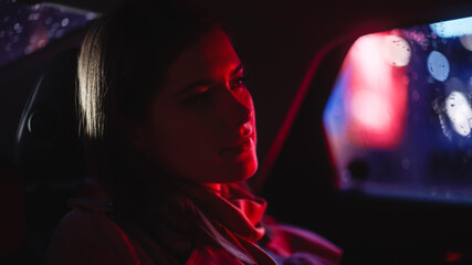 Close Up Portrait of a Female Commuting Home in a Backseat of a Taxi at Night. Beautiful Woman Passenger Looking Out of Window while in a Car in City Street with Working Neon Signs.