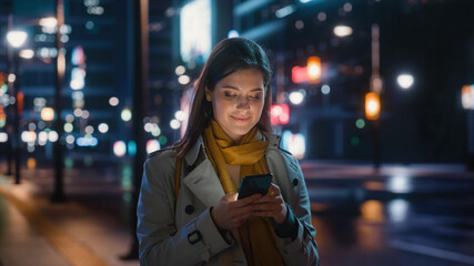 Fototapeta premium Portrait of a Beautiful Woman in Trench Coat Walking in a Modern City Street with Neon Lights at Night. Attractive Female Using Smartphone and Looking Around the Urban Cinematic Environment.