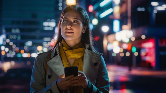 Portrait Of A Beautiful Woman In Trench Coat Walking In A Modern City Street With Neon Lights At Night. Attractive Female Using Smartphone And Looking Around The Urban Cinematic Environment.