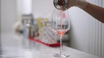 Close-up pouring pink orange cocktail from shaker in drinking glass in slow motion. Slim female Caucasian hand pours liquid alcoholic beverage in pub bar indoors. Alcohol industry concept