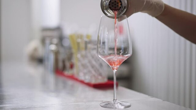 Close-up pouring pink orange cocktail from shaker in drinking glass in slow motion. Slim female Caucasian hand pours liquid alcoholic beverage in pub bar indoors. Alcohol industry concept