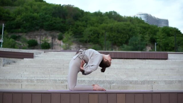 Wide shot of young flexible woman performing bending body backward exercises, various gymnastic and yoga exercises sitting on yoga mat. Sporty yogini female practicing backbend outdoors in city park.
