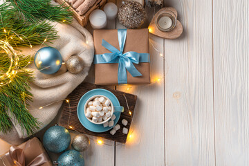 A cup of coffee with marshmallows, Christmas decorations and a gift on a light background illuminated by a burning garland.