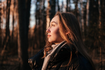 Smiling young woman in coat and scarf standing in autumn forest. Woman looking at the sunset on trees background and breathing fresh air. Person in nature. Having fun outdoors.