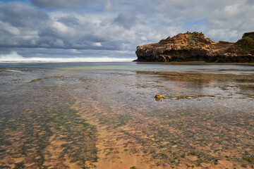 Long exposure of waves washing into a sandy beach shore, with a dragon shaped rock in the background and cloudy blue sky on the horizon, in Victoria, Australia.