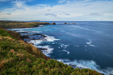 Overlooking a scenic view of a beautiful rocky coastline with waves breaking, deep blue sea in the background and cloudy blue sky on the horizon, photographed at Phillip Island, Australia.