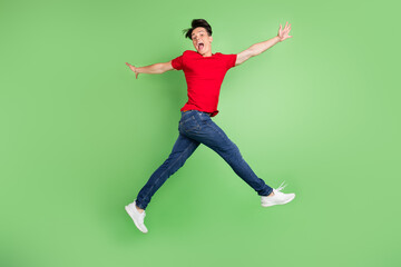 Fototapeta na wymiar Photo of lucky funky young man wear red t-shirt smiling jumping high walking isolated green color background