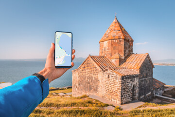 Naklejka premium a hand with phone with maps app for navigating to attractions. At the background - Lake Sevan and the famous Armenian monastery Sevanavank