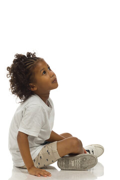 Small black boy in shorts and shirt is sitting on a floor and looking up. Side view. Full length, isolated.