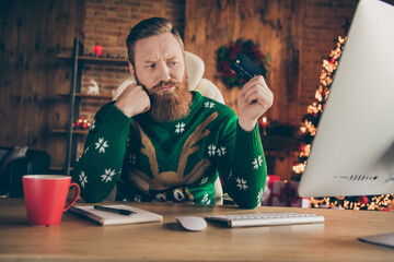 Photo of focused doubtful man sit desk hand chin think look debit card work pc wear deer pullover...