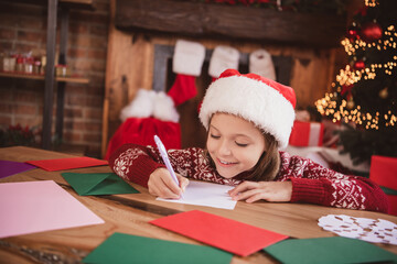 Photo of charming pretty schoolgirl dressed red sweater writing christmas congratulations smiling...