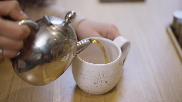 Close-up pouring green herbal tea from teapot in cup. Unrecognizable young Caucasian woman pouring healthful detox organic hot drink in cafe restaurant indoors. Slow motion