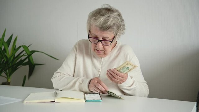 An elderly upset woman in glasses sits in a bright room at home and counts banknotes in her hands, reads text in a notebook. The concept of poverty, limited budget and small pension and old people