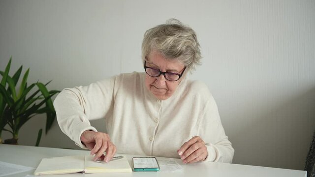 An elderly woman in room puts on glasses, writes in a notebook with a pen and calculates taxes and expenses on a calculator in a smartphone. Concept of budget calculation and pensioners' finances