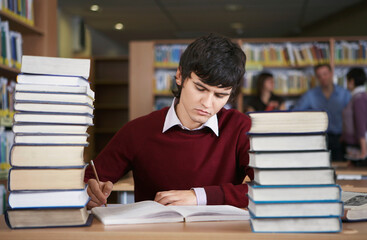 Student writing at desk with stacks of books in library