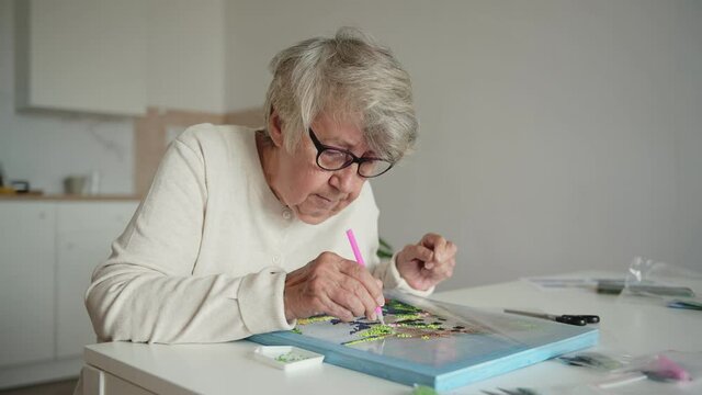 Elderly grey-haired woman in warm woolen jacket and glasses spends retirement time laying out diamond embroidery at table