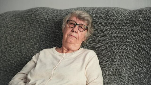 Upset elderly lonely gray-haired woman with wrinkled face wearing glasses and white cardigan cries taking off spectacles leaning on sofa back