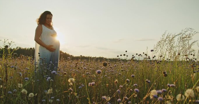Pregnant woman with a big belly feeling happy outdoors. Adult expecting mother stroking belly on nature.  Middle age white pregnant woman touching her belly while sunset. Woman pregnancy concept.