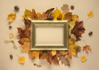 A golden frame surrounded by autumn leaves on a beige (cream) background. Autumn composition, decoration.