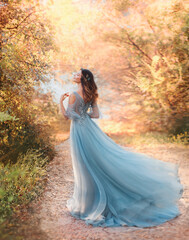 happy fairy woman princess in light summer blue dress standing in autumn park orange foliage tree....