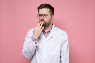 Caucasian doctor in a white coat pensively and anxiously looks to the side, holding hand to face. Pink background.