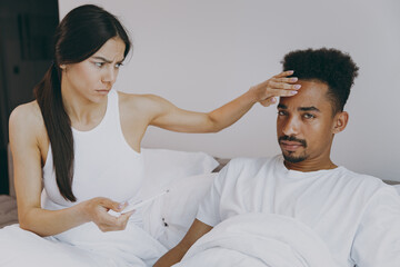 Fototapeta premium Sick ill tired disturbed young couple two family in casual white clothes sit in bed woman measure temperature to man hold thermometer spend time together in bedroom lounge home own room house wake up.