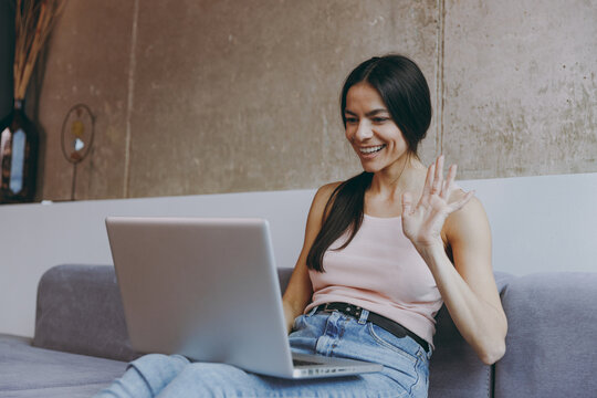 Smiling cheerful young latin woman 20s in casual clothes sit on sofa hold use talk work on laptop pc computer get video call spend time in living room home in own house be lost in reverie good day