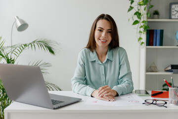 Young friendly fun smiling happy successful employee business woman in casual blue shirt sit work at workplace white desk with laptop pc computer at modern office indoors. Achievement career concept.