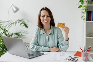 Young fun smiling successful employee business woman in blue shirt hold in hand credit bank card...