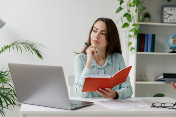 Young minded pensive thoughtful employee business woman in blue shirt writes in notebook sit work at workplace white desk with laptop pc computer at modern office indoors Achievement career concept.