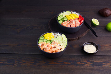 Poke bowl with salmon,shrimps, avocado, rice, cucumber, mango, pepper and sesame seeds, soy sauce on dark wooden background. 