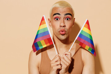 Young shocked amazed happy fun blond latin gay man with make up in beige tank shirt holding rainbow...