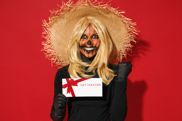 Young overjoyed woman with Halloween makeup mask wears straw hat black scarecrow costume hold gift certificate coupon voucher card for store do winner gesture isolated on plain red background studio