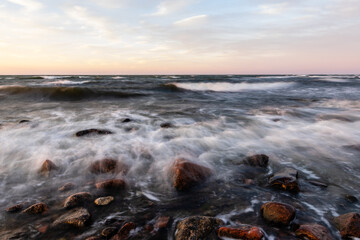 Fototapeta premium Long exposure photo of waves reaching to rocky coast during dusk. Whitecaps waves are reaching to shore during sunset. Motion of ripples and coastal rocks at sundown