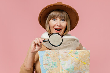 Traveler tourist amazed mature elderly senior lady woman 55 years old wears brown shirt hat scarf hold examine map with magnifying glass isolated on plain pastel light pink background studio portrait.