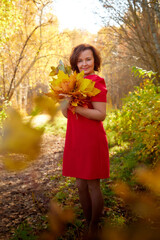 Naklejka premium Portrait of middle-aged woman with dark curly hair and in red dress in autumn park with yellow and green leaves