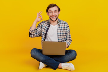 Full size photo of a happy young man holding laptop computer show okey sign while sitting on a floor isolated over yellow color background