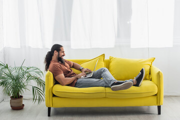 side view of hispanic man sitting on yellow couch at home and using laptop