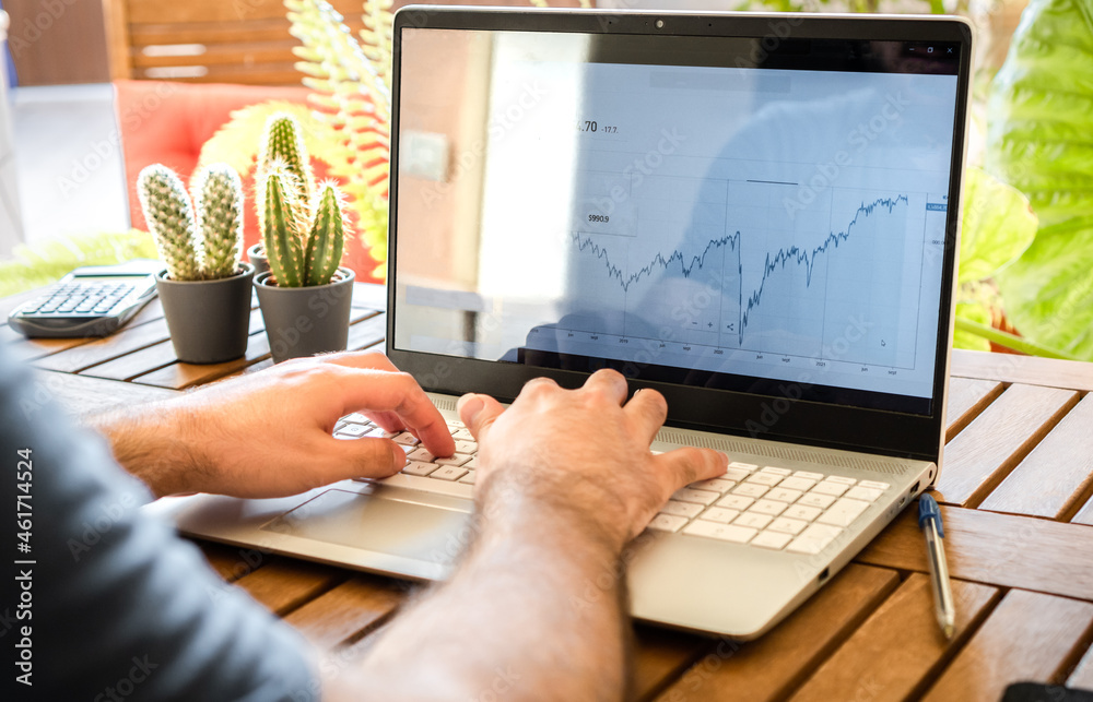 Wall mural young man sitting at table investing in stock market with laptop during work remotely in an outdoor 